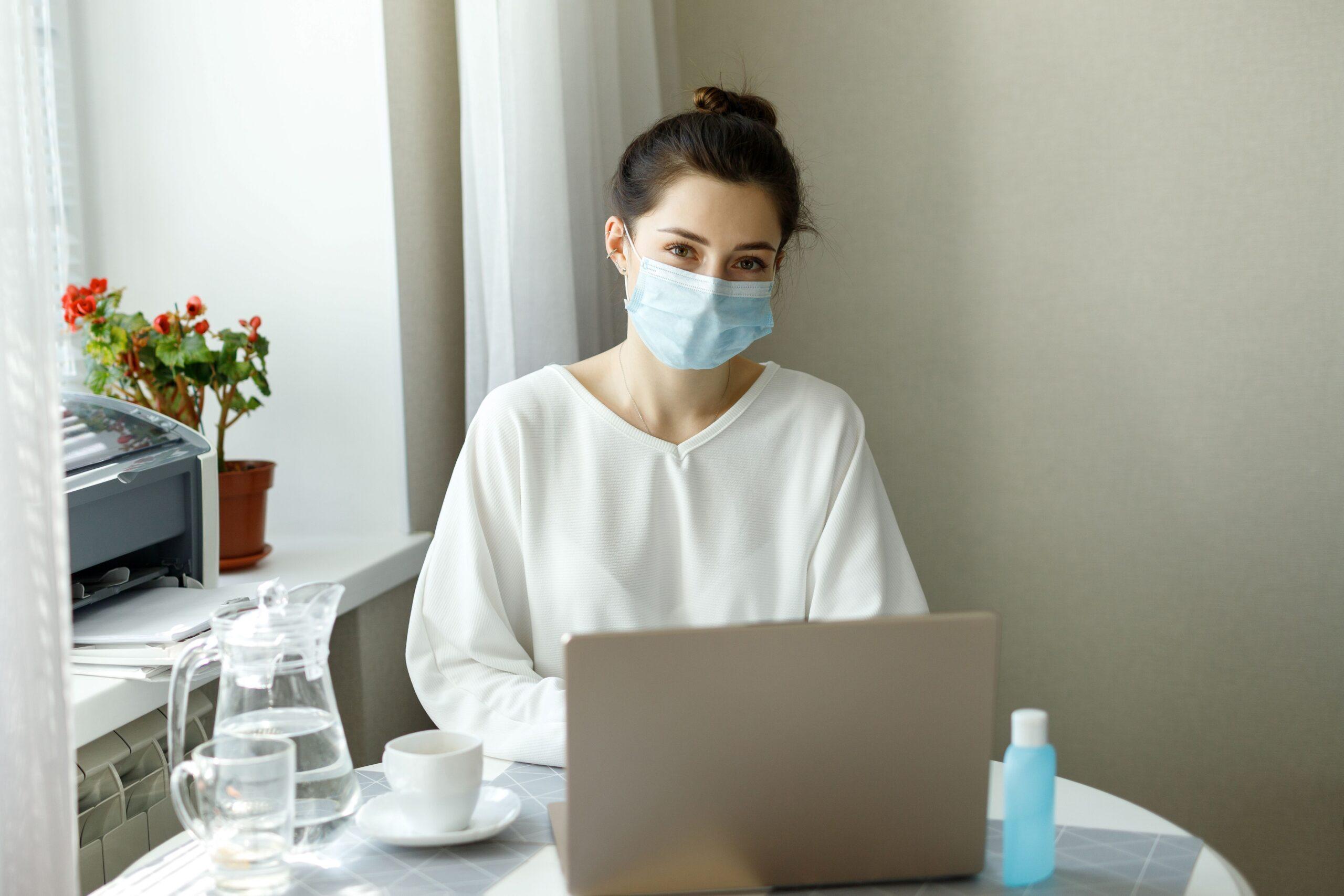 Frau mit Maske am Laptop, Desinfektionsmittel auf dem Tisch